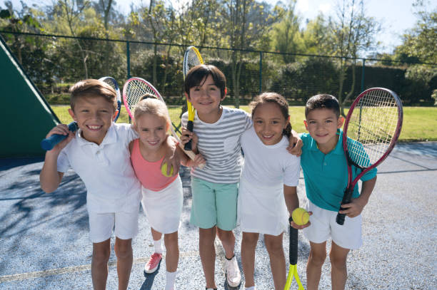 Group of children at the tennis court embracing each other and holding their rackets looking at camera smiling very happy